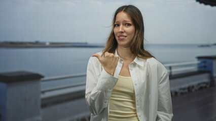 Woman points thumb over her shoulder on seaside promenade street, visible hand and thumb with uneasy facial expression; doubt hesitation refusal.
