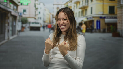 Fototapeta premium Young hispanic woman clenches fists and throws head back in a joyful shout on a crowded city street, smiling with closed eyes; triumph celebration.