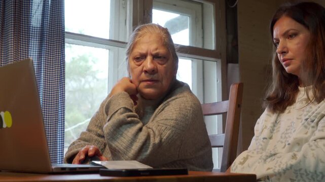 Two women, an older mother and her daughter, look with serious and concerned expressions at a laptop in a wooden home interior. They are reviewing information at a table, possibly financial documents