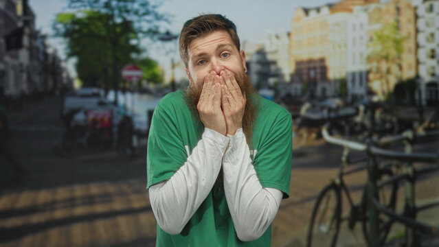 Volunteer man in green shirt covers mouth with hands on city street beside canal and parked bicycles; surprise.