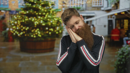 Young caucasian man with beard rests cheek on folded hands in sleeping gesture on urban street; serenity.