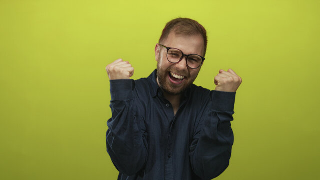 Man with clenched fists and glasses laughing, wearing a navy shirt, in a lime green studio; joy celebration success.