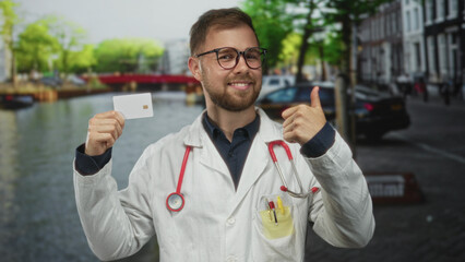 Man doctor in white coat with stethoscope holds card and points finger on an amsterdam street canal, wearing glasses and smiling; trust health confidence.