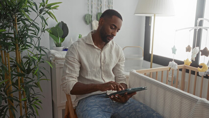 Man taps tablet beside baby crib in a nursery room, hand touching screen while seated on chair by window; calm focus.