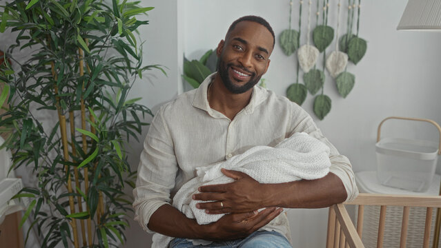 Young african american man gently cradling newborn baby with bare hands in bright nursery room beside wooden crib; fatherhood bonding serenity.
