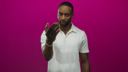 Young african american man pointing finger at camera, visible hand and finger, wearing white polo in magenta studio setting; challenge.