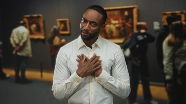 Man with hands crossed on chest standing in a museum building wearing white shirt and looking at camera while visitors view paintings; contemplation empathy. - Powered by Adobe