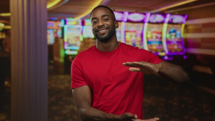 Young african american man in red shirt gesturing with hands as if holding invisible prize at casino slot machines; luck playful chance.