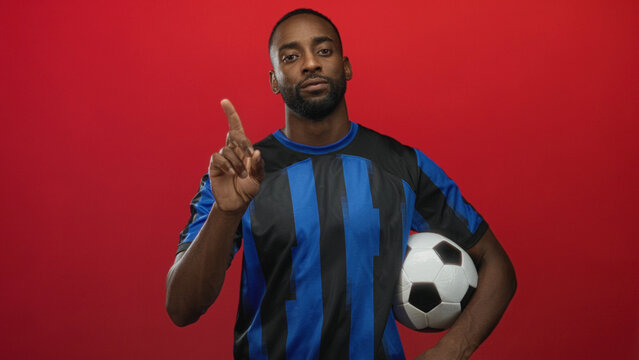 Man holding soccer ball and pointing finger in studio against red backdrop, wearing blue striped jersey; confidence focus.
