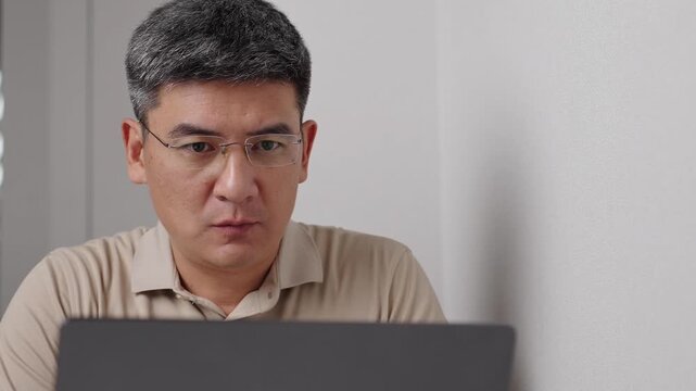 Shocked man sits at sleek desk concentrating on his laptop in bright, modern workspace. Asian man browsing online with surprised facial expression. Businessman stunned with unexpected news.