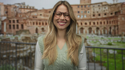 Blonde woman with round glasses framing her smiling face with hands amid roman forum ruins; travel joy wonder.
