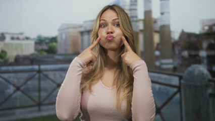Fototapeta premium Blonde woman wearing a pink top points finger to puffed cheeks against rome street railing with blurred ruins; playfulness humor silliness.