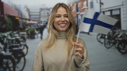 Blonde woman smiling while holding finnish flag on busy city street; national pride patriotism support.