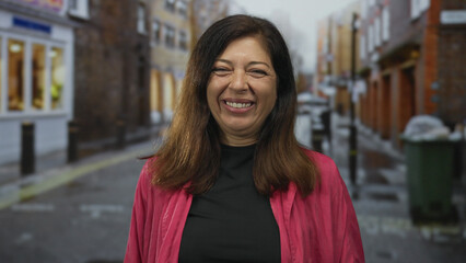 Middle age hispanic woman wearing pink jacket sticks out tongue on rainy urban street; playfulness.