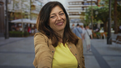 Woman in casual brown jacket with warm smile extends hand toward camera on bustling street; connection.