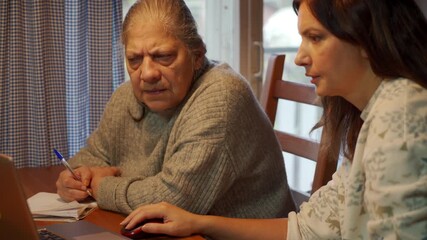 A concerned elderly woman takes notes in a notebook with a pen while her daughter uses a laptop mouse. The women are focused on an online task, possibly for paperwork, learning, or bill paying.