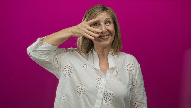 Middle aged woman covers right eye with hand over white blouse against vibrant pink studio wall; playful curiosity.