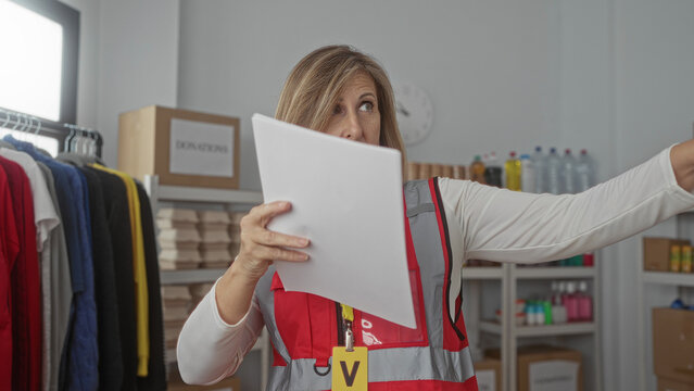 Woman volunteer in red vest holds list paper with finger pointing in building donation center; compassion. - Powered by Adobe