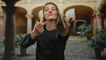 Young blonde woman shows middle fingers with scowl toward viewer in old building courtyard under...
