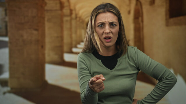 Young blonde woman points finger toward camera with hand on hip in building corridor of old town, confrontational forward stance; anger confrontation.