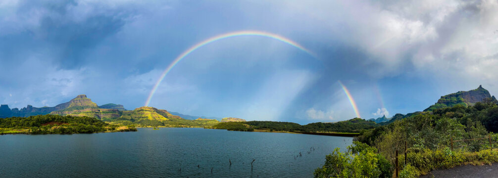 Panoramic view of Ghatghar Dam in Bhandardara featuring a vibrant rainbow over lush green mountains and a serene reservoir after rainfall.

