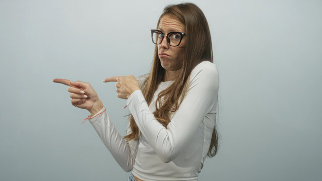 Woman wearing glasses and long brown hair points both index fingers across the frame in a studio against a pale blue wall; skepticism doubt uncertainty.
