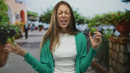Woman holding lime near face and grimacing with closed eyes and open mouth on street, wearing green...