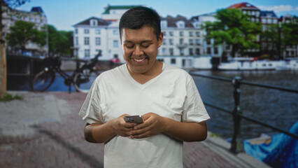 Man smiling and holding smartphone with both hands while texting on street by amsterdam canal and railing; happiness.