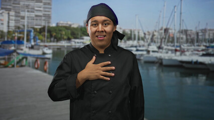 Hispanic man chef in black uniform with hand on chest at marina by boats next to building; pride...
