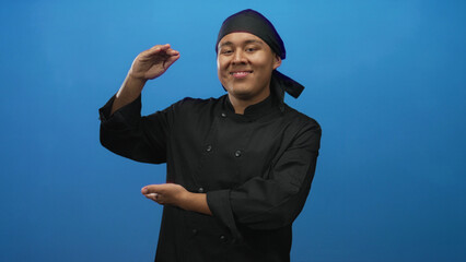 Man hispanic chef in black uniform smiling and framing empty space with hands in studio blue; pride confidence.