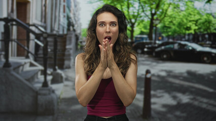 Young hispanic woman holds fingers near chin with open mouth on bustling city street; surprise.