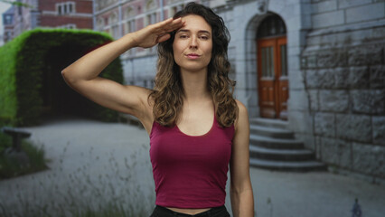 Woman in maroon tank top with long wavy hair salutes on street by stone stairs with focused expression; pride.