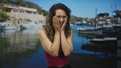 Woman presses her hands to her cheeks at building waterfront dock while smiling broadly and closing...