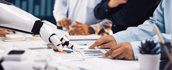 A robot hand interacts with human colleagues during a business meeting. They analyze various charts and graphs, showcasing collaboration in a modern office setting.Noogenesis
