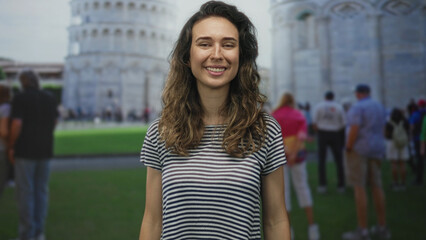Young hispanic woman lifts open palms at leaning tower building in crowded piazza under bright sunlight; joy travel.