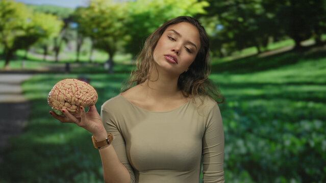 Young hispanic woman holds a brain model above her shoulder with extended arm in green forest; mental fatigue.