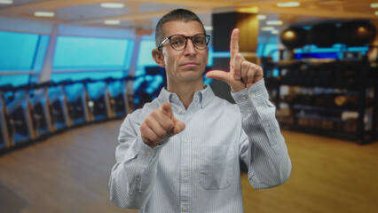 Man in glasses making loser gesture and pointing at camera in a brightly lit indoor gymnasium with a row of fitness equipment and modern design.
