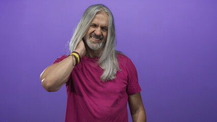 Man holding neck with hand in studio with purple backdrop, long gray hair and winced expression; discomfort reflection.