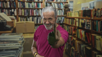 Man with hoary long hair shows middle finger and clenched fist in library building among bookshelves and stacked books, wearing rings and bracelets; defiance.