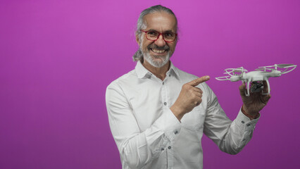 Man points finger at drone while holding a quadcopter in a purple studio, wearing white shirt and red glasses; curiosity innovation.
