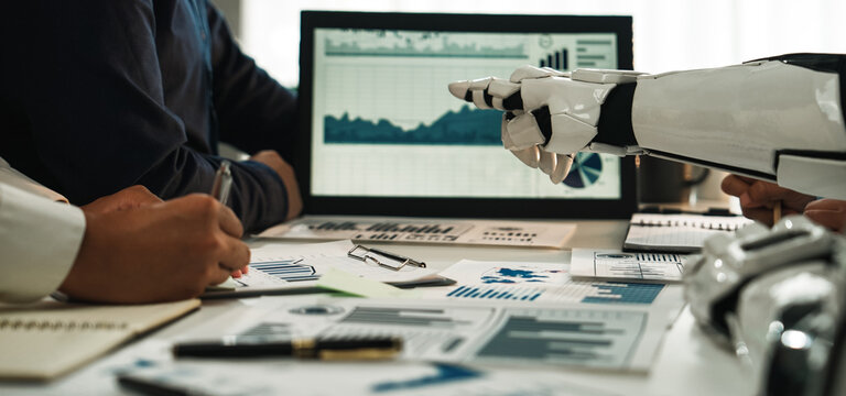 A modern workspace featuring a robot hand pointing at data analysis charts during a professional meeting. The scene symbolizes innovation and collaboration in business.Noogenesis