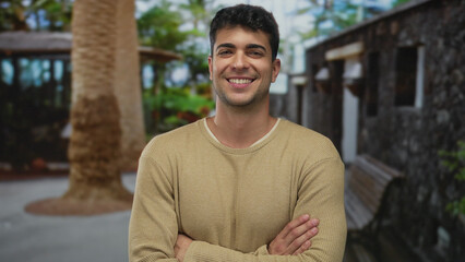 Man holds arms across chest and smiles outdoors showing young hispanic style over crossed urban benches with a calm grin portrait.