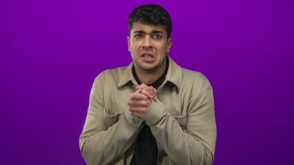 Man grasps hands in a gesture against a background, hispanic young male anxious purple studio scene.