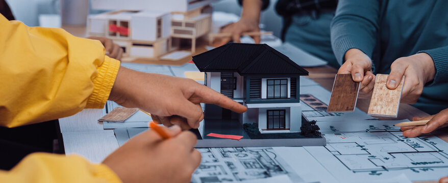 A team collaborates on an architectural project, showcasing a model house, blueprints, and material samples. The scene illustrates creativity and teamwork in design. SACTR