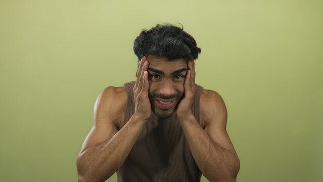 Man with muscular arms, tense facial expression, and open palms pressed to temples against green studio wall; confusion.