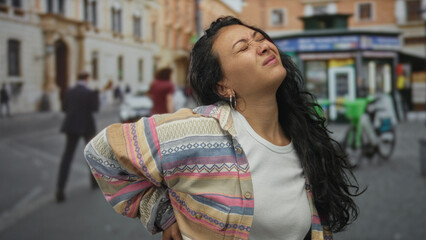 Fototapeta premium Woman holding lower back with backache and grimacing on street in front of a kiosk and bicycles; pain struggle.