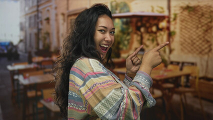 Fototapeta premium Woman points fingers on street terrace of a restaurant while smiling and gesturing with hands at camera; playful confidence.