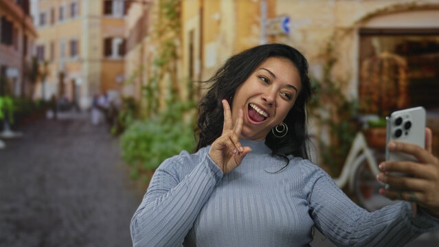 Woman holding smartphone for selfie on street in a sunlit european alley, smiling and flashing a peace sign while gesturing with hand; joy connection. - Powered by Adobe