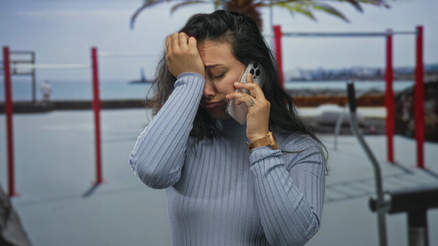 Woman holding smartphone to ear with hand on forehead near outdoor gym bars in street; phone call sadness.