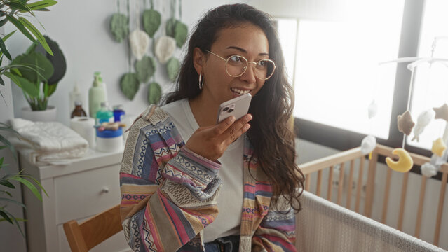 Woman holding smartphone by wooden crib in baby room, speaking into phone while seated on chair near changing table; calm caregiving.